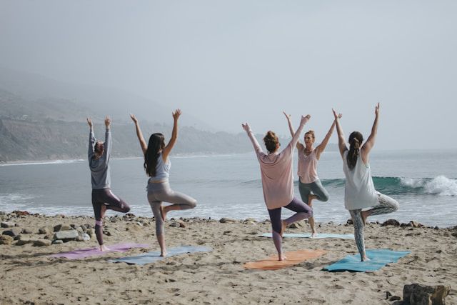 Five women at the beach, standing on one leg, with their arms in the air